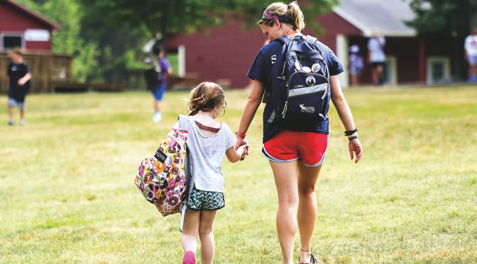 A camp counselor walks hand-in-hand with a young camper near the cabins