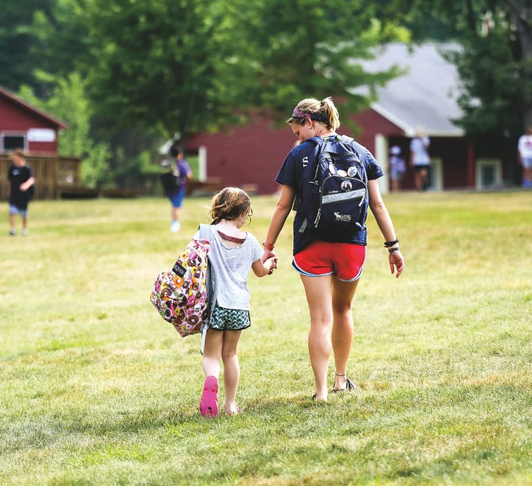 A camp counselor walks hand-in-hand with a young camper near the cabins