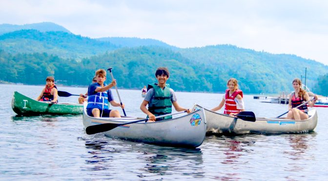 A canoe group paddles together on a calm lake at summer camp