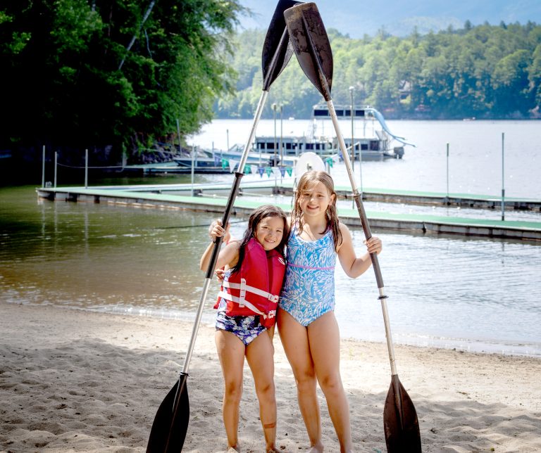 Two young campers with linked arms and paddles crossed overhead, smiling by the lake